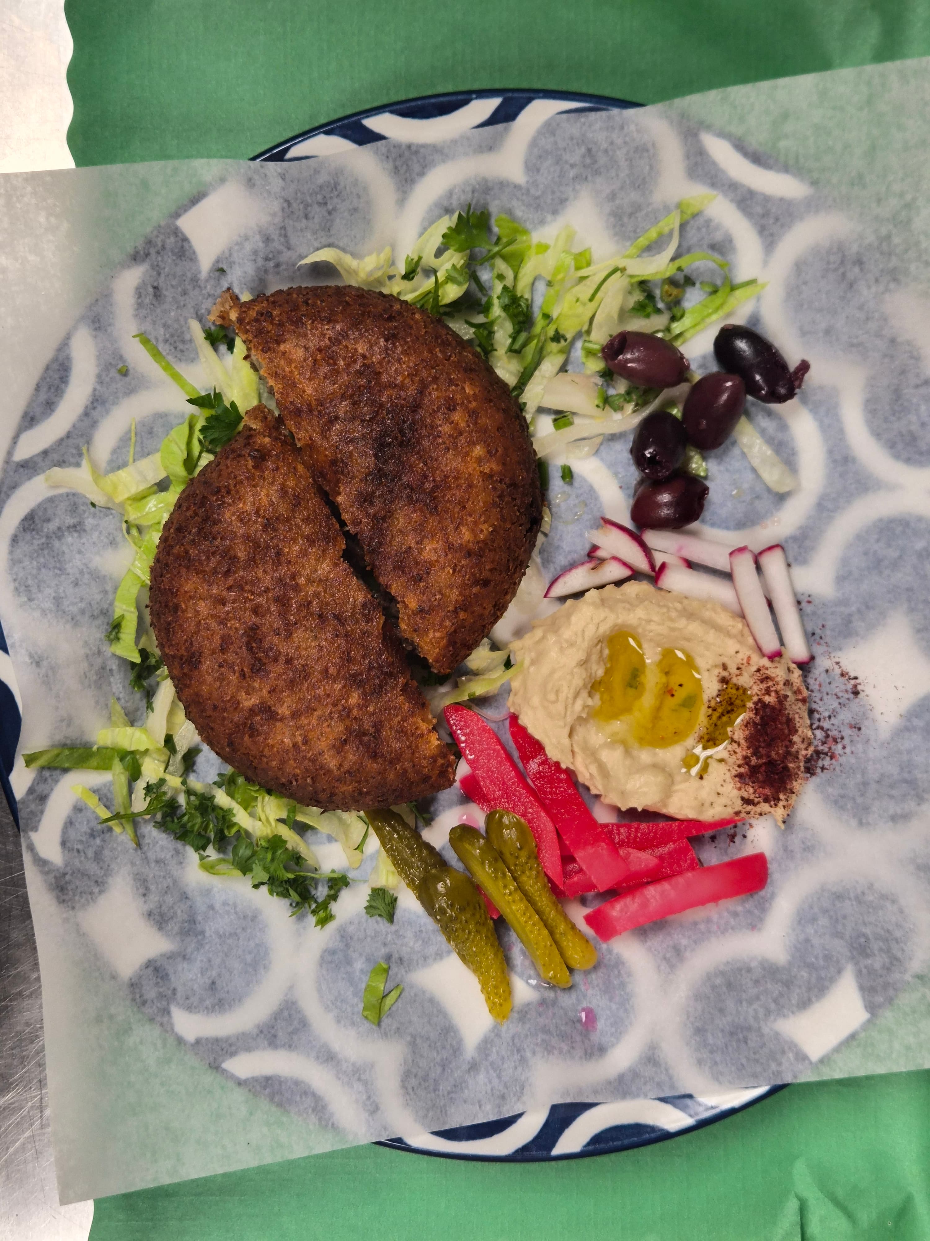 Fried kibbeh halves on lettuce with hummus, olives, and pickled vegetables on a patterned plate.