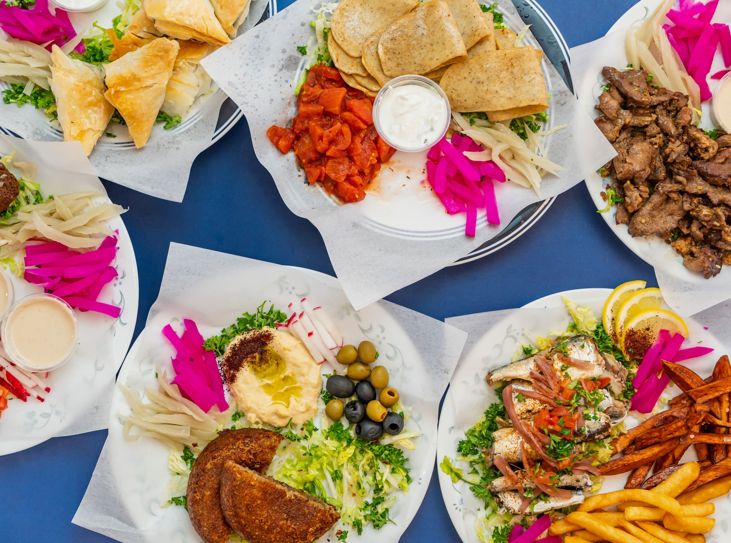 Assorted Mediterranean plates featuring hummus, falafel, grilled meats, and colorful pickled vegetables on blue background.