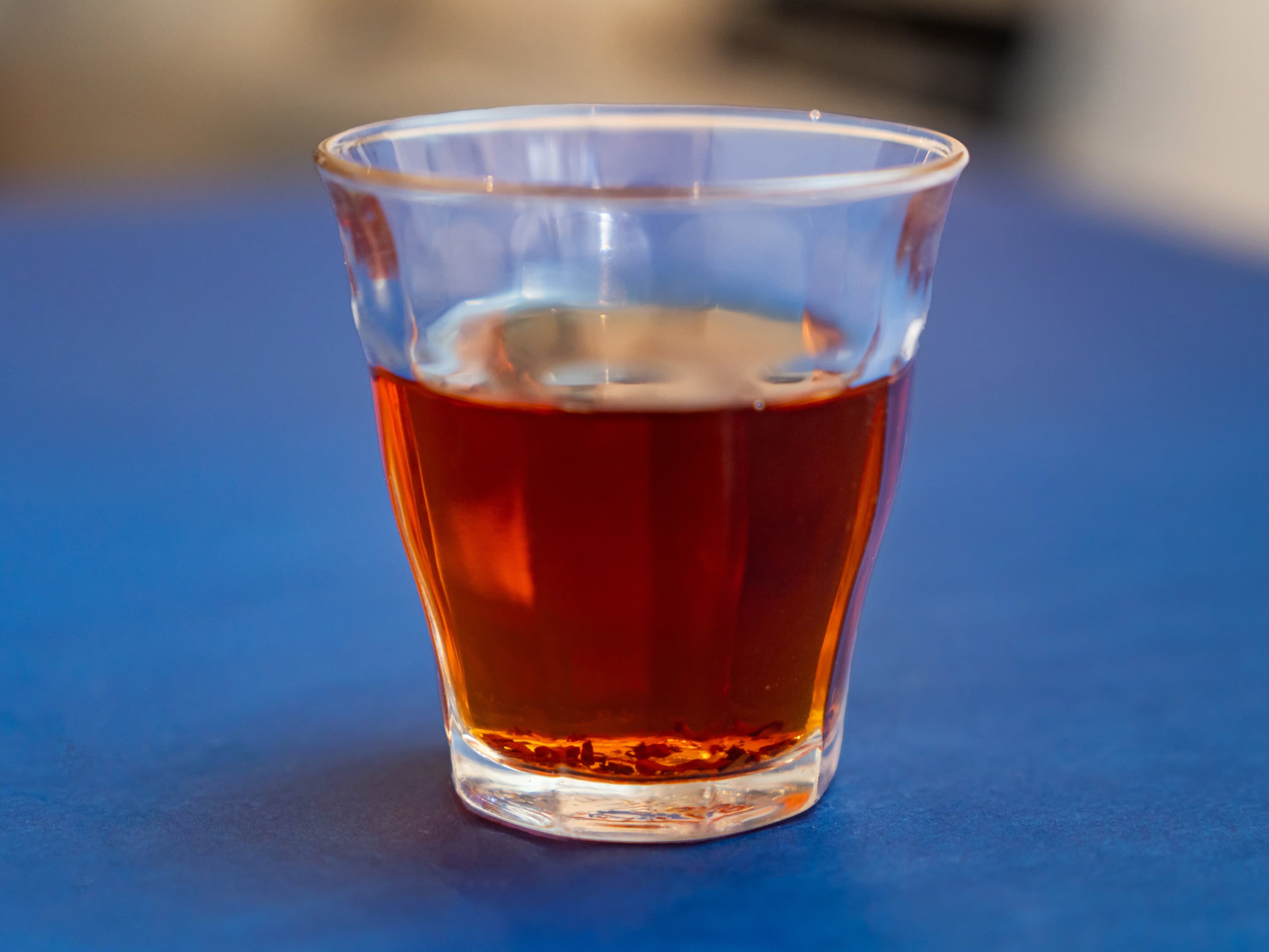 Small clear glass of amber tea with loose leaves on a solid blue background.