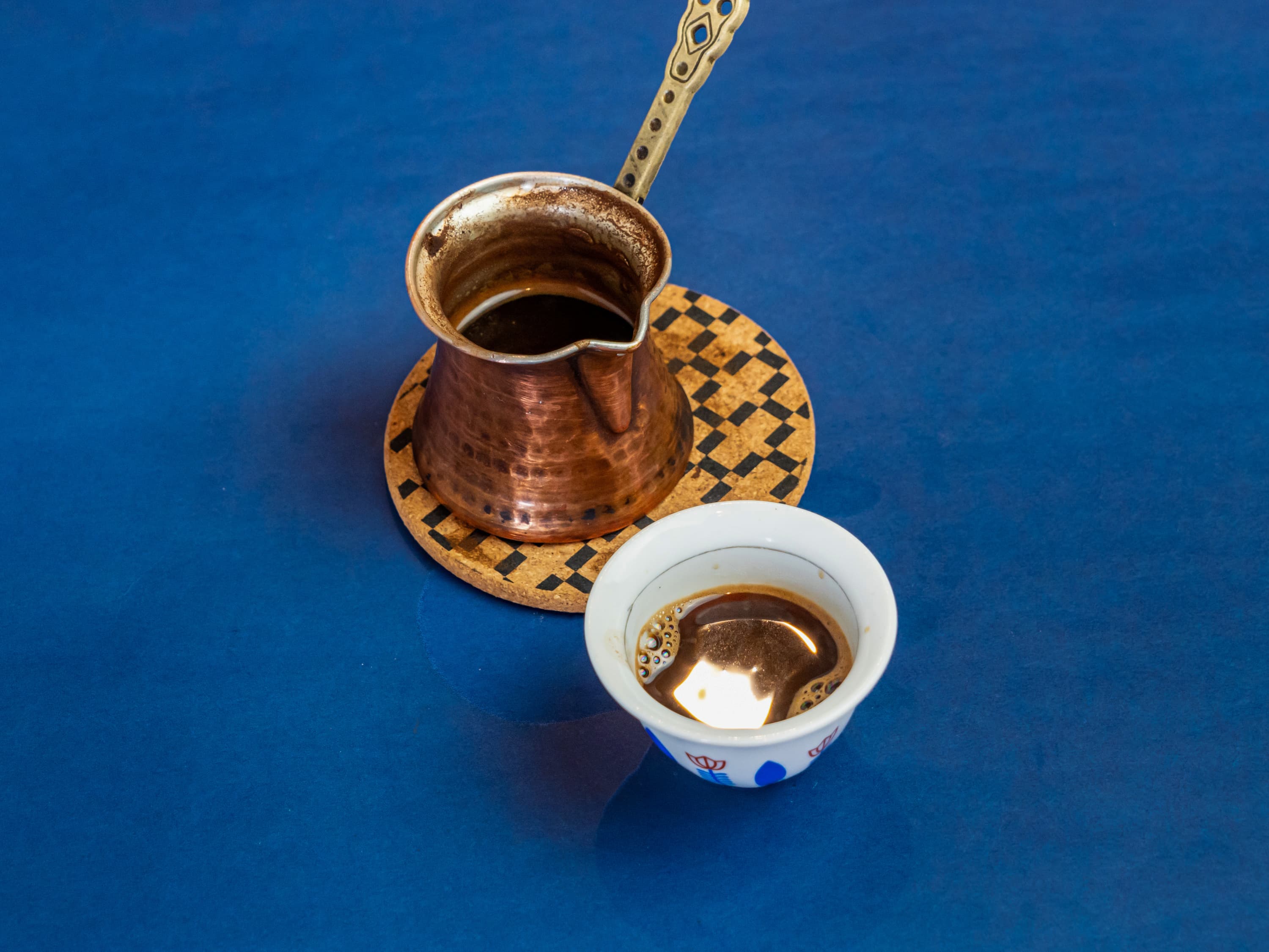 Copper Turkish coffee pot and a small white cup of coffee on blue background.