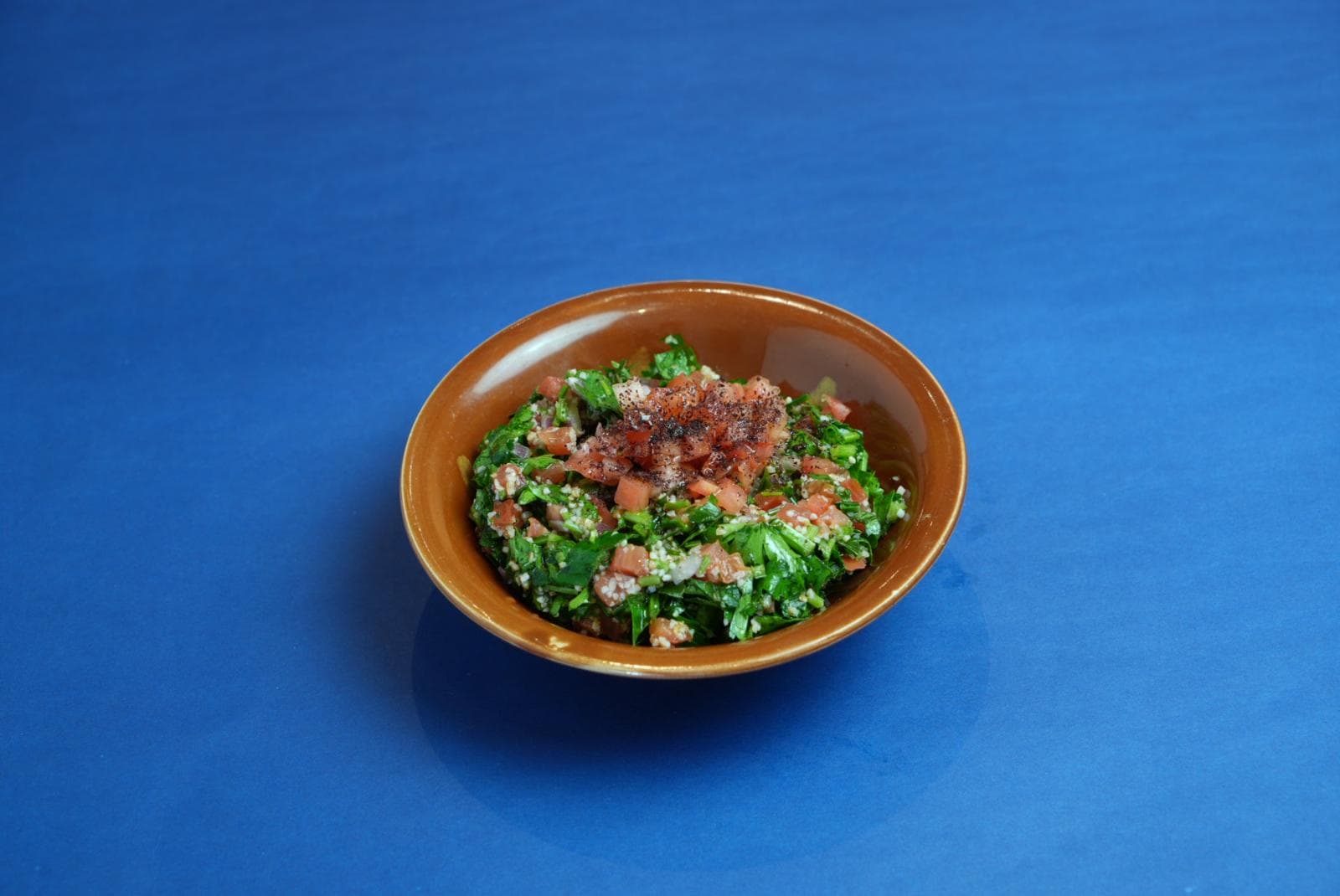 Tabbouleh salad with parsley, tomatoes, and bulgur in a brown bowl on blue background.