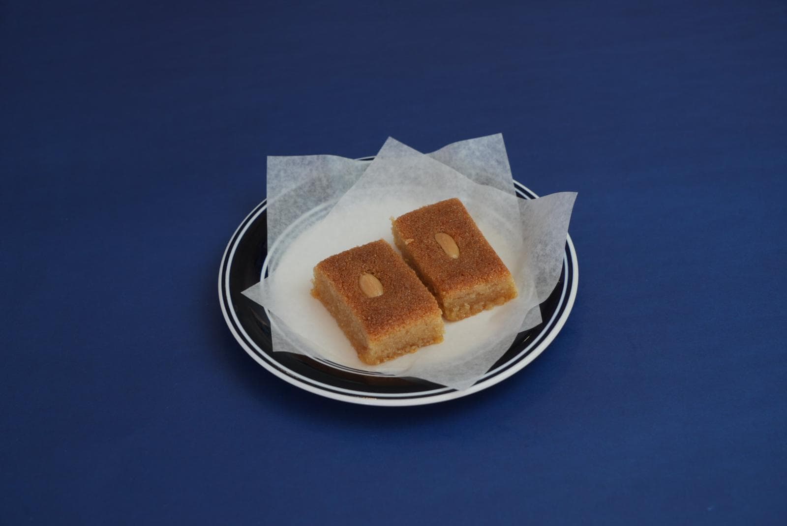 Two golden semolina cake squares topped with almonds on a striped plate over blue background.