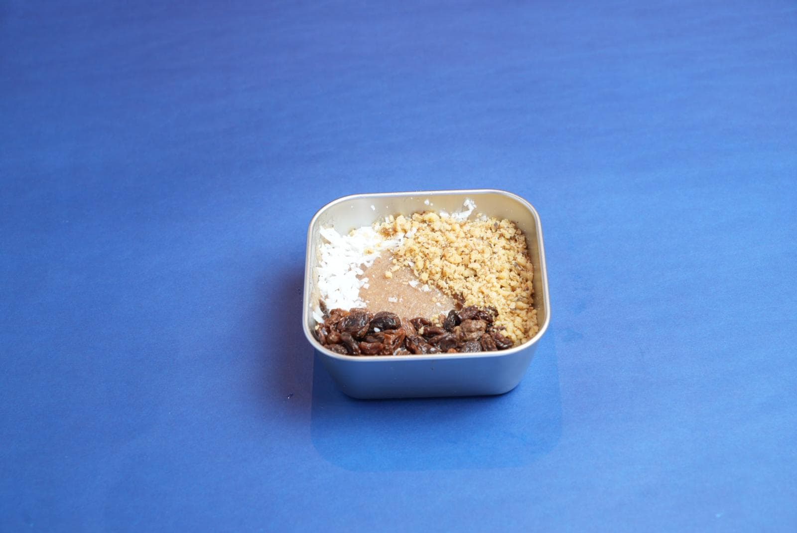 Square metal bowl containing brown porridge topped with coconut, nuts, and raisins on blue surface.