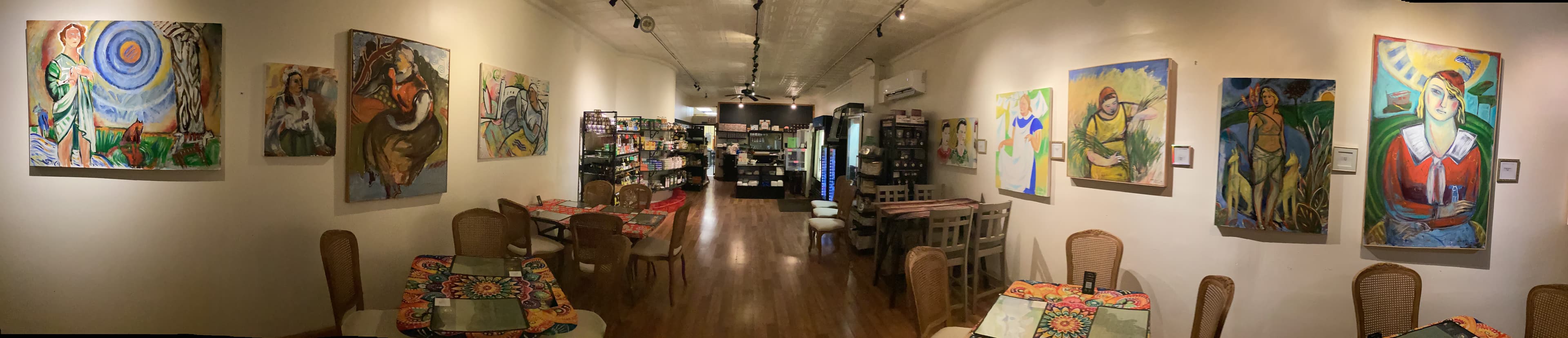 Panoramic view of a cafe interior featuring colorful paintings, wooden floors, and patterned tablecloths.