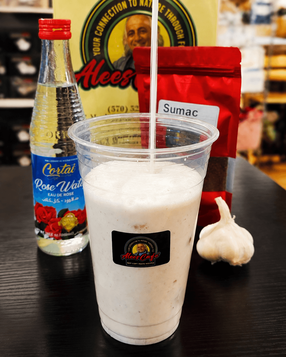 Frothy white drink in a plastic cup alongside rose water, sumac, and fresh garlic.